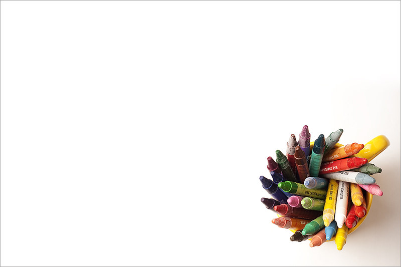 Crayons in approximately 25 different colors arranged in a yellow mug and photographed from above, placed in the lower right corner of a white rectangle