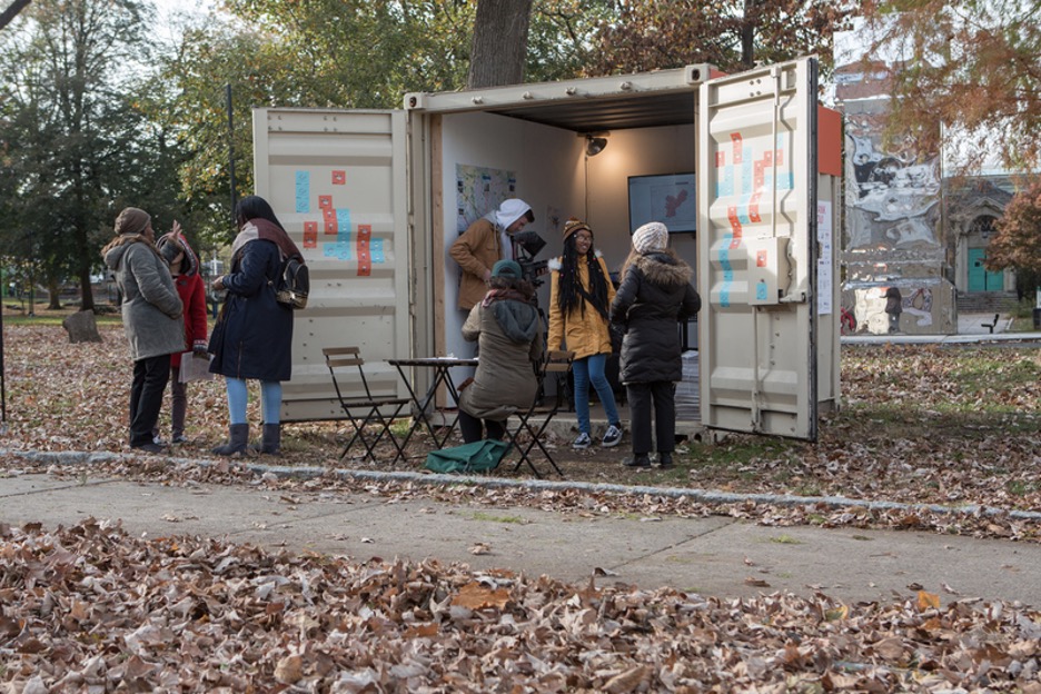 Image of several people standing near a pop-up art gallery in a metal shipping container near a sidewalk. Fall leaves cover the ground.